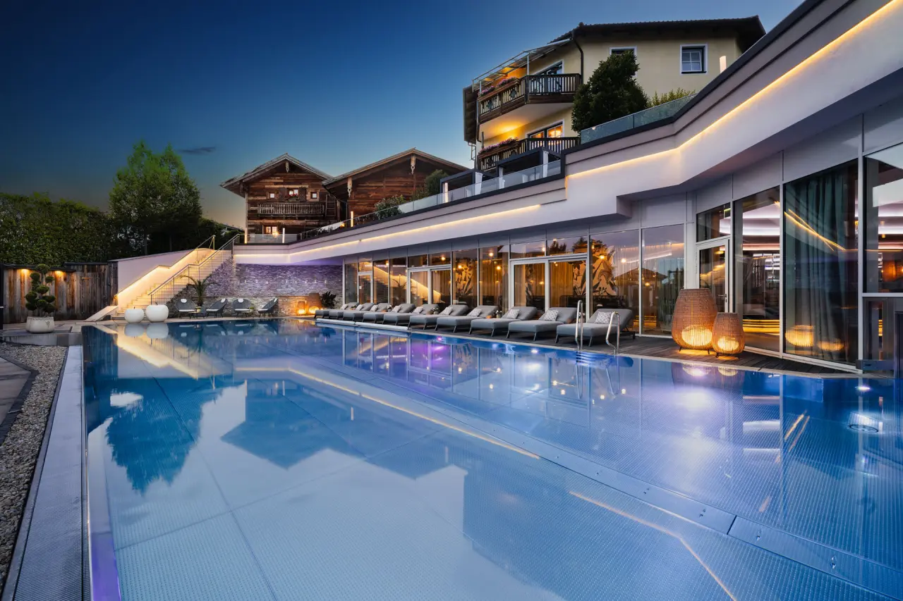 Luxuriöser Außenpool des 5-Sterne Wellness- & Sporthotels Jagdhof bei Dämmerung mit beleuchteter Fassade und Liegen.