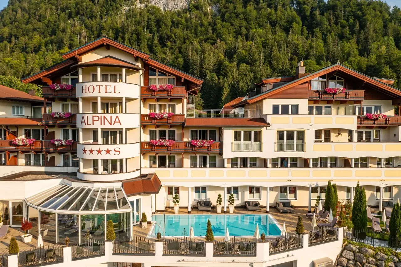 Aussenansicht des Hotel Alpina mit sonnigem Aussenpool und Liegebereich vor bewaldeter Bergkulisse.