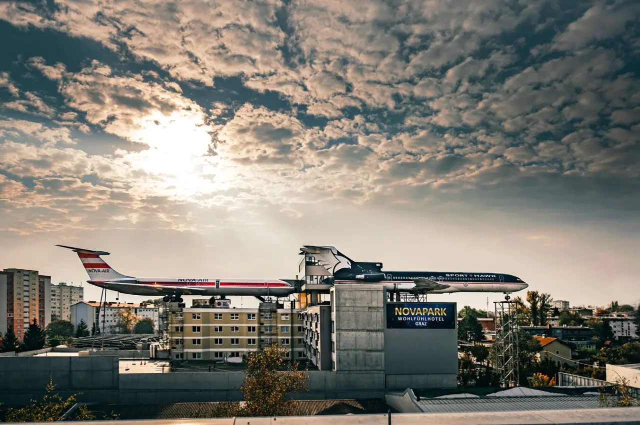 Novapark Flugzeughotel Graz: Spektakuläre Aussenansicht mit zwei Flugzeugen auf dem Dach unter dramatischem Himmel.