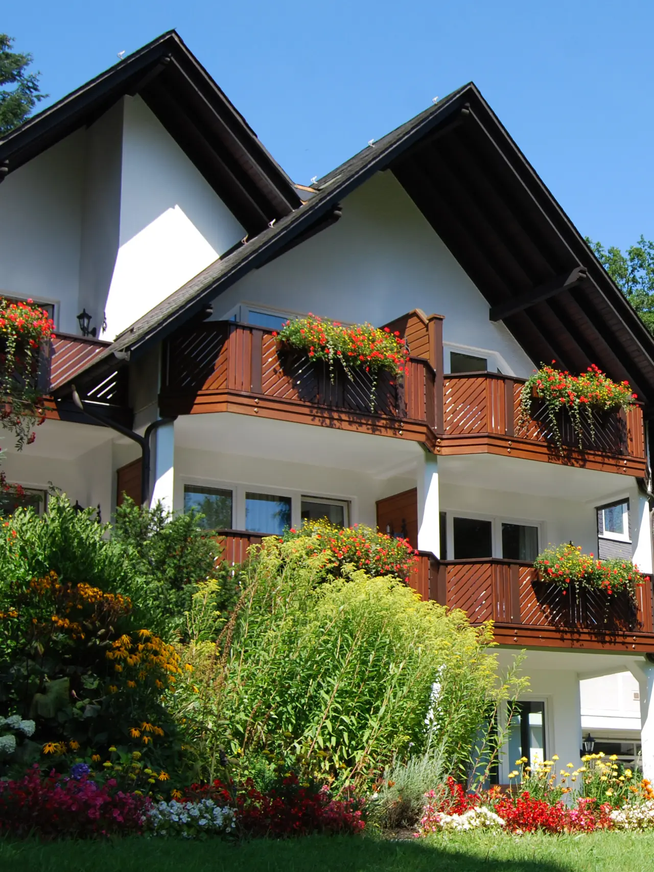 Aussenansicht des Hotel Haus Hilmeke mit blühenden Balkonen und gepflegtem Garten unter blauem Himmel.