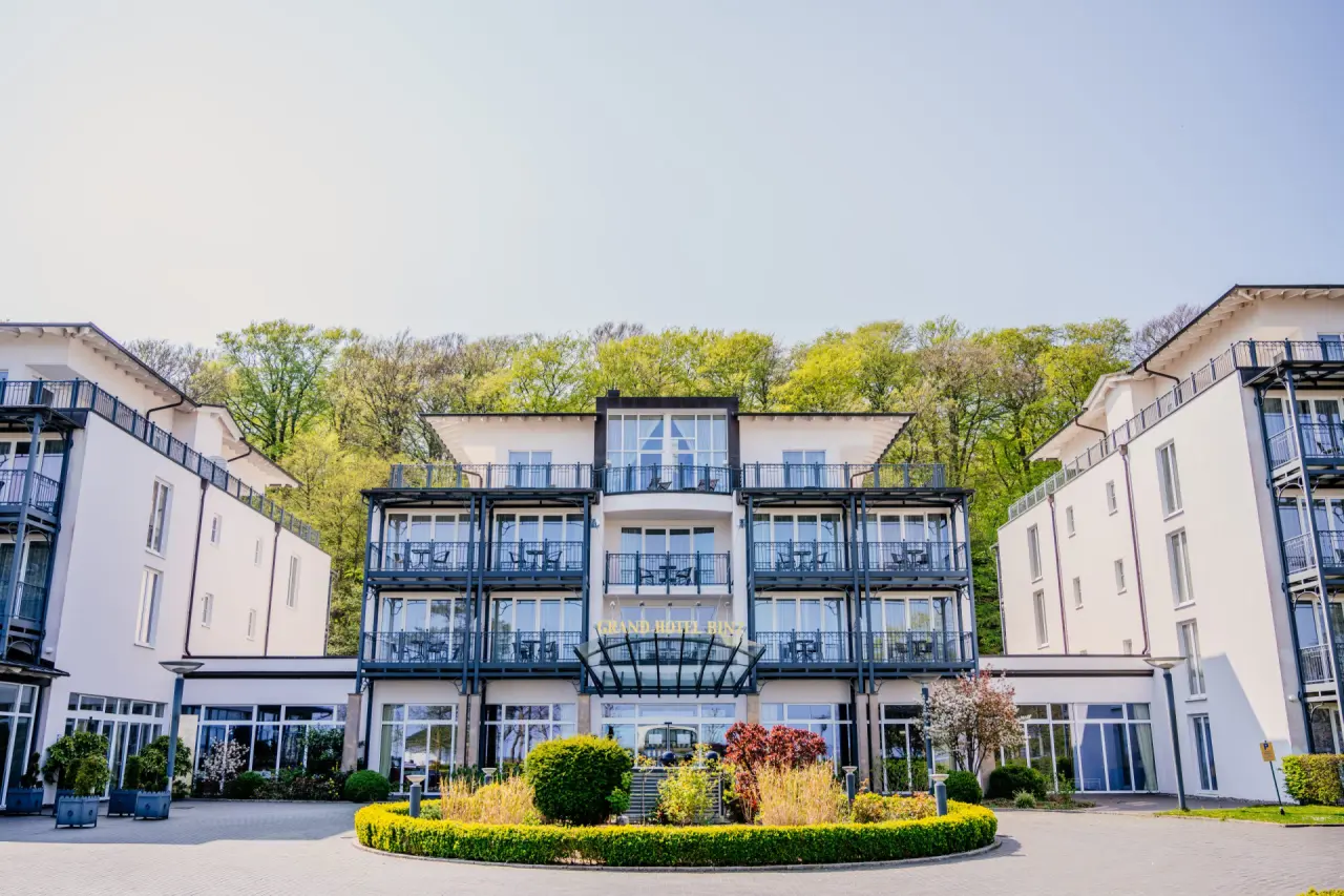 Elegante Aussenansicht des Grand Hotel Binz mit modernen Balkonen und gepflegter Zufahrt, umgeben von Baeumen.