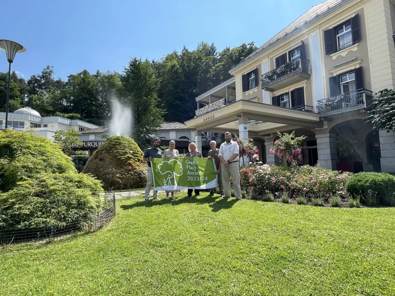 Aussenansicht Hotel Warmbaderhof mit Green Flag Award Banner und Mitarbeitern. Grüner Garten mit Brunnen und historischem Gebäude.