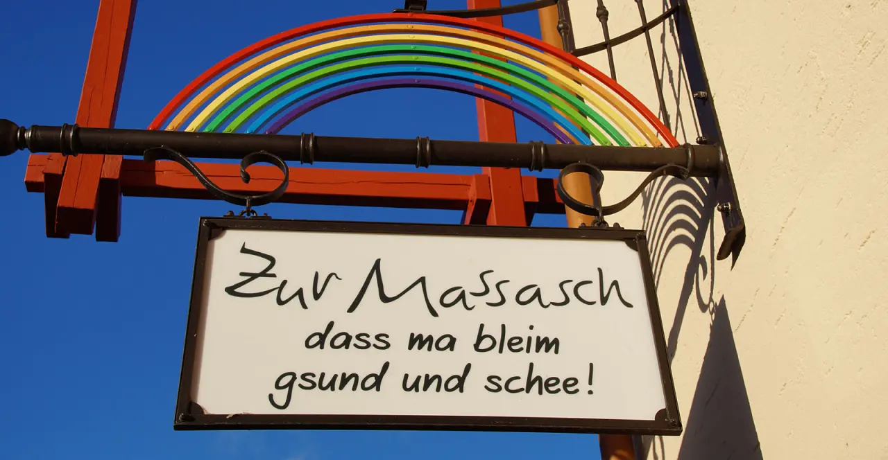 Hängendes Holzschild mit Regenbogen und bayerischem Spruch 'Zur Massasch dass ma bleim gsund und schee!' am Hotel Zum Kramerwirt.