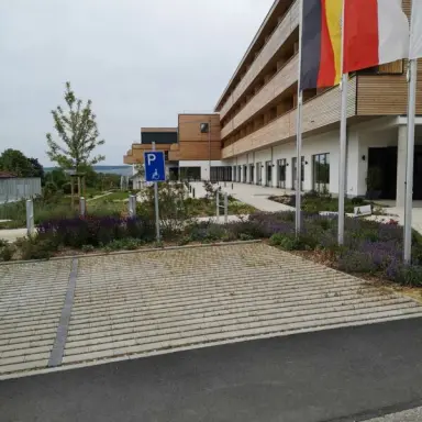 Aussenansicht des sonnenhotel WEINGUT RÖMMERTH mit moderner Holzfassade, Parkplatz und Blumenbeeten unter bewölktem Himmel.