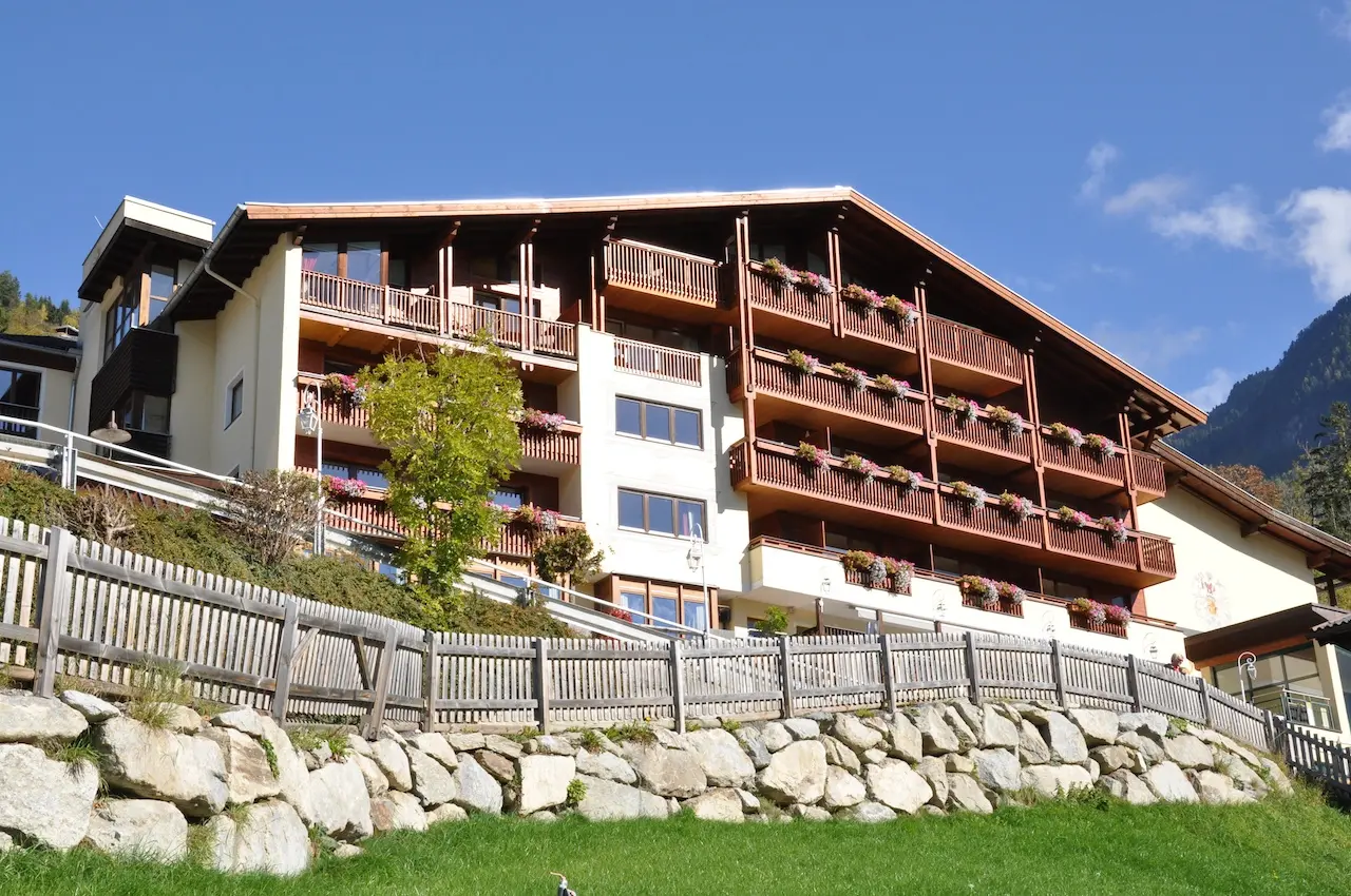 Aussenansicht des Wellnesshotels Jerzner Hof im alpinen Stil mit Holzbalkonen, Blumenkästen und Bergkulisse unter blauem Himmel.
