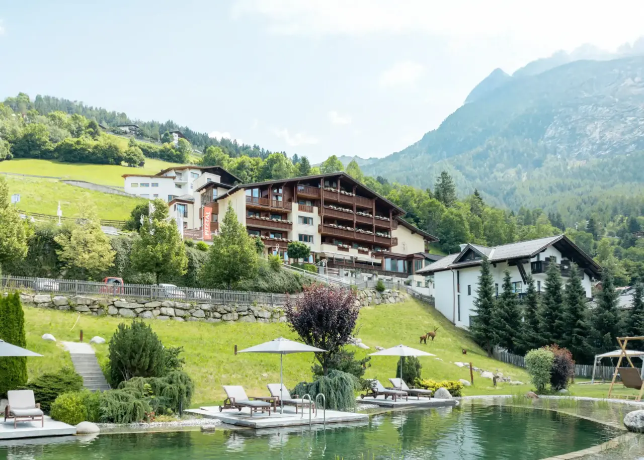 Naturbadeteich mit Liegewiese und dem Hotel Jerzner Hof im Hintergrund, umgeben von grünen Bergen im Pitztal.