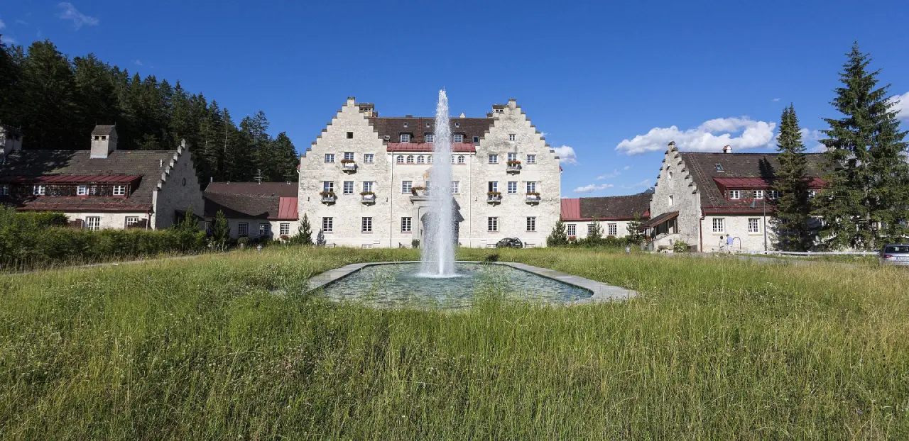 Historisches Hotel Das Kranzbach mit Brunnen und Wald im Hintergrund unter blauem Himmel.