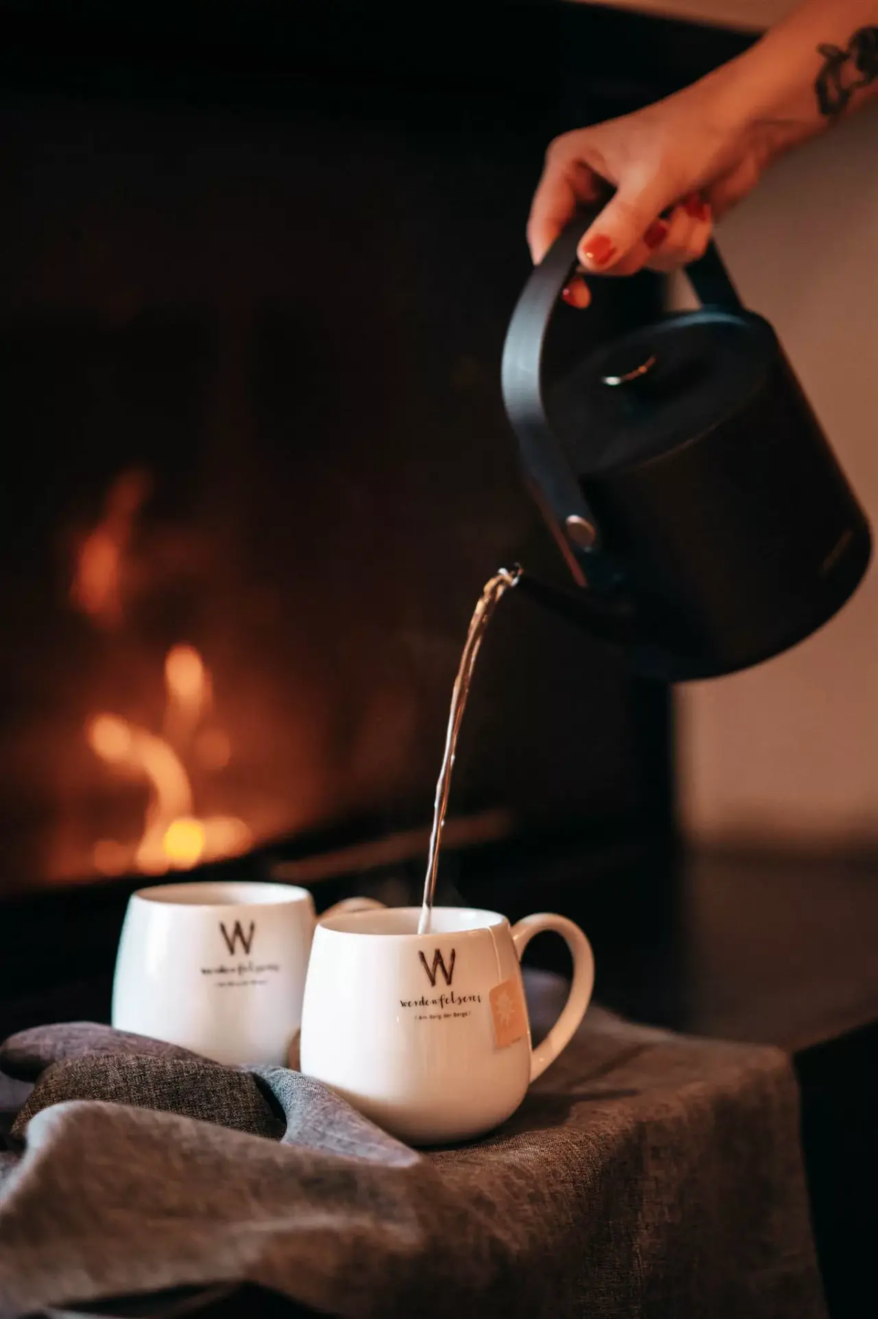 Gemütlicher Abend am Kaminfeuer im Hotel WERDENFELSEREI. Eine Hand gießt dampfenden Tee in eine Tasse mit Hotel-Logo.