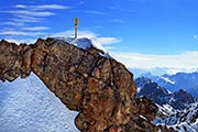 Gipfelkreuz auf der Zugspitze mit verschneiten Felsen und blauem Himmel, nahe dem Hotel Zugspitze.