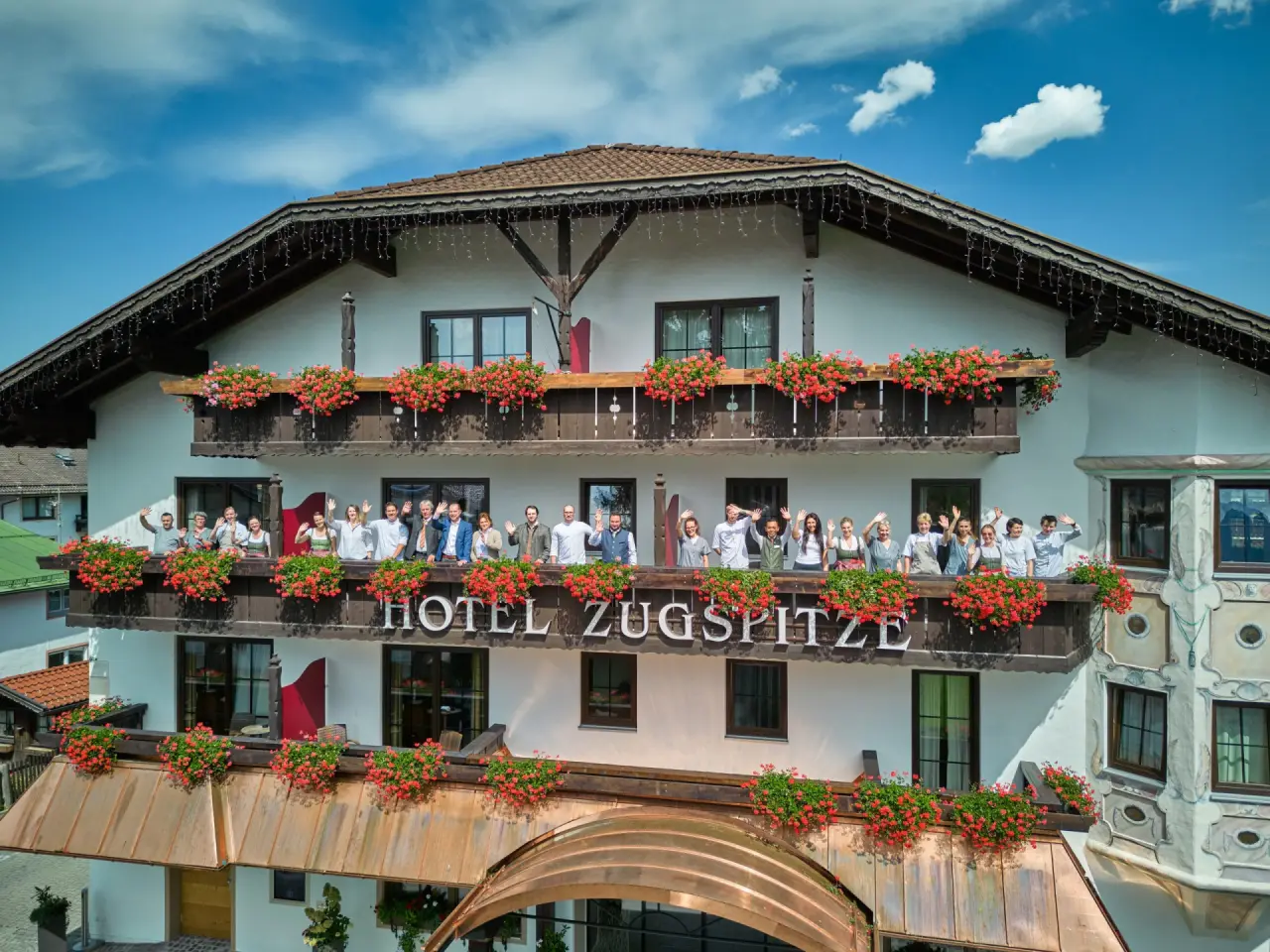Aussenansicht des Hotel Zugspitze mit blühenden Balkonen und winkendem Personal unter blauem Himmel.