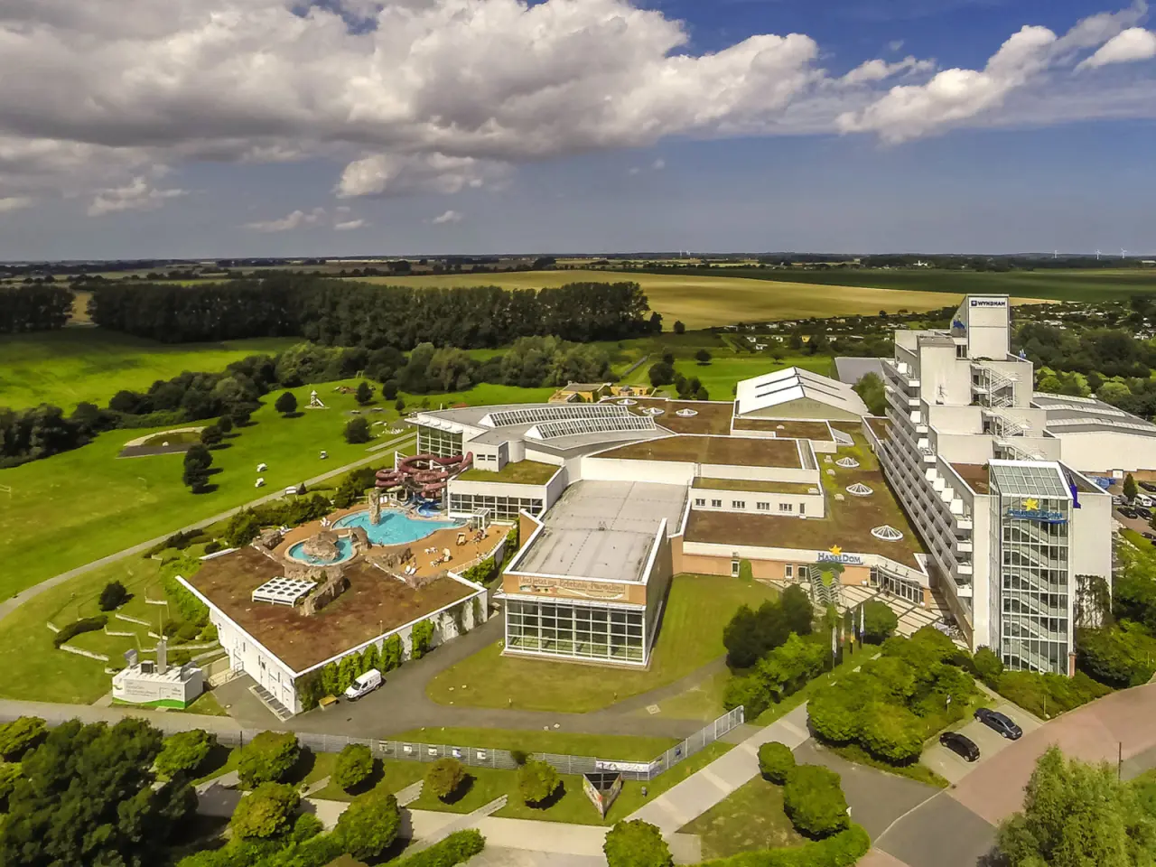 Luftaufnahme des Wyndham Hotel Stralsund HanseDom mit großem Außenpool, Wasserrutschen und weitläufiger Grünanlage unter blauem Himmel.