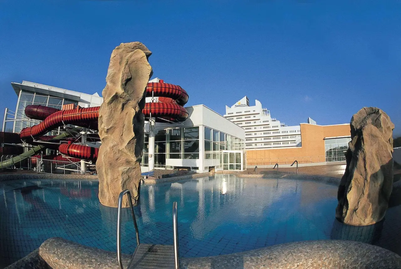 Aussenpool mit Wasserrutschen und Hotelgebäude des Wyndham Hotel Stralsund HanseDom unter blauem Himmel.