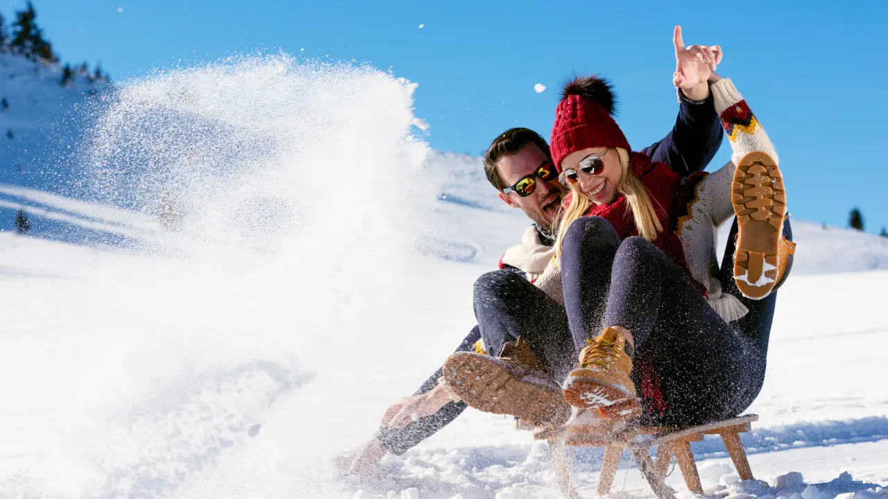 Fröhliches Paar beim Rodeln im Schnee vor winterlicher Bergkulisse am Hotel Salnerhof.
