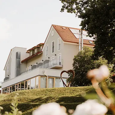 Aussenansicht des Hotels Das Eisenberg mit moderner Architektur, rotem Dach und Herz-Skulptur im gruenen Garten.