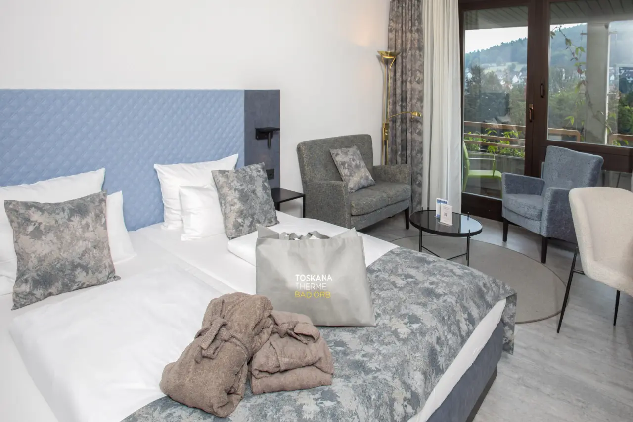 Geräumiges Hotelzimmer im Hotel an der Therme Bad Orb mit Doppelbett, Sitzbereich und Balkon mit Waldblick.