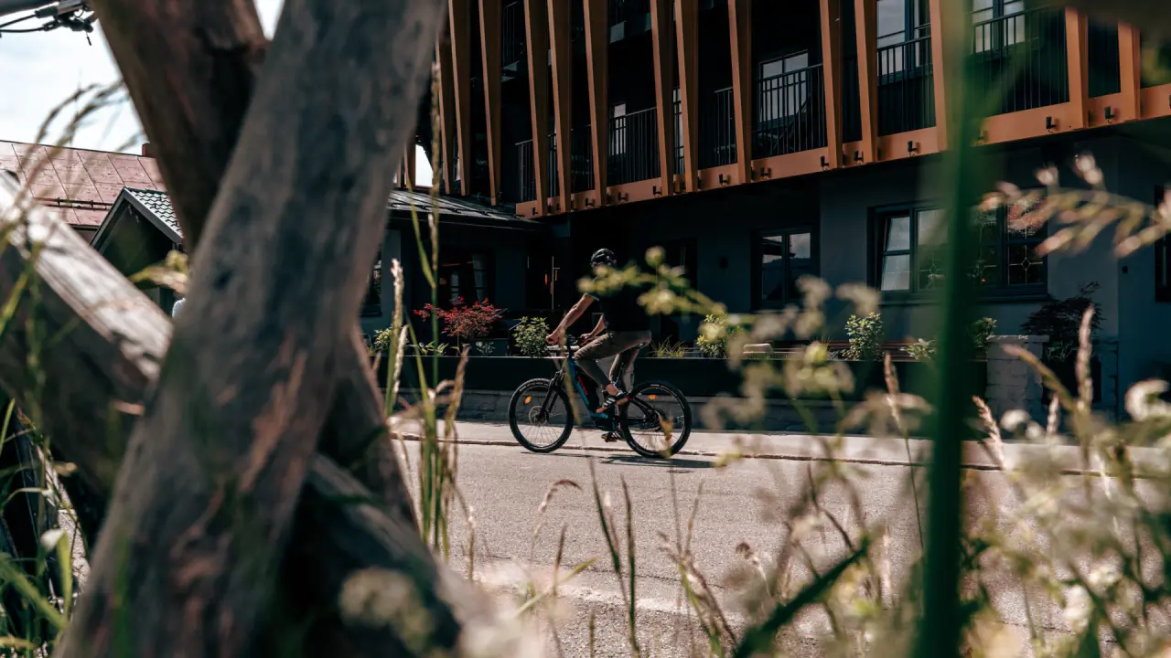 Aussenansicht des Hotel Alpenkönig mit moderner Holzfassade und einem Radfahrer auf der Strasse, durch Bäume verdeckt.