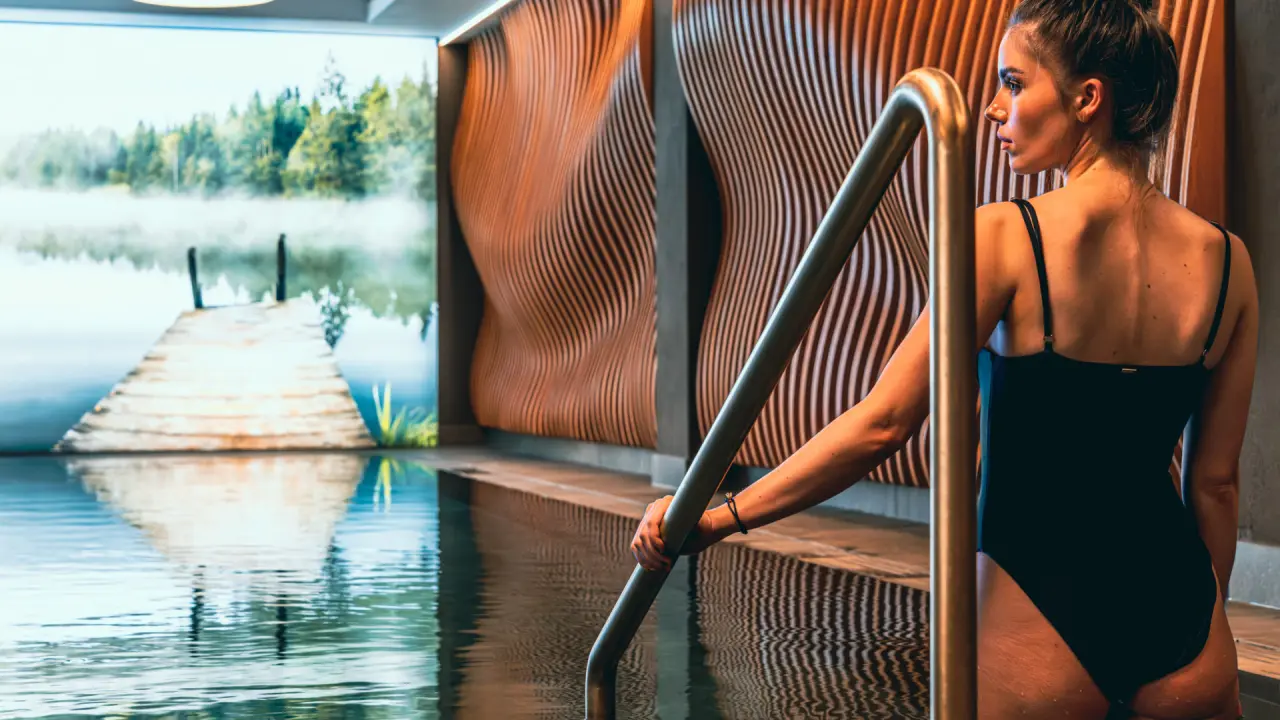 Moderne Poollandschaft im Hotel Alpenkönig mit stilvoller Holzverkleidung und beruhigendem Seemural. Eine Frau entspannt am Beckenrand.