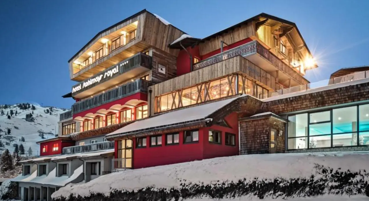 Beleuchtetes Hotel Kohlmayr Royal im Winter bei Dämmerung, umgeben von Schnee und Berglandschaft, mit sichtbarem Poolbereich.