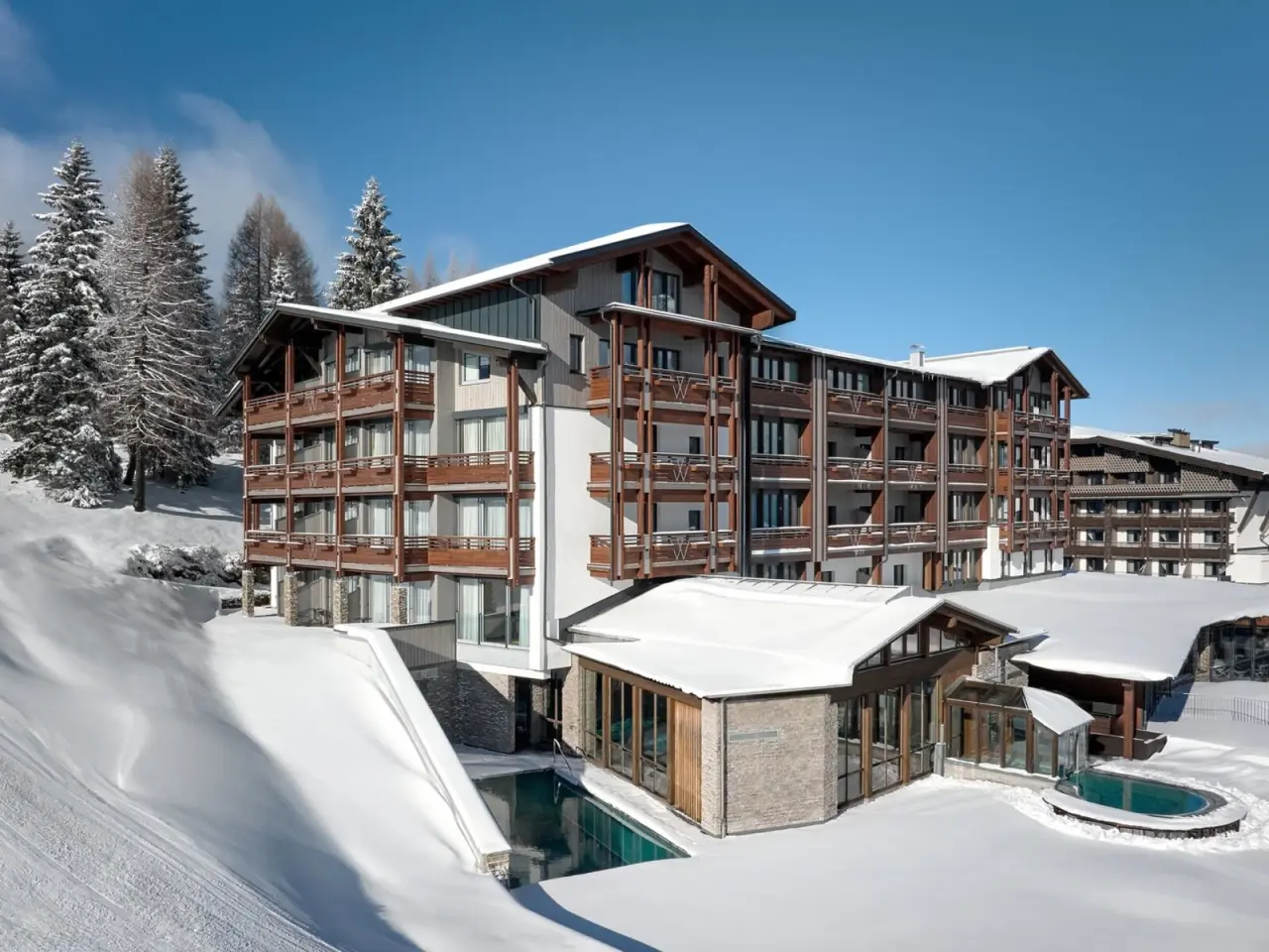 Aussenansicht des Hotel & Spa Wulfenia im Winter mit schneebedeckter Landschaft und beheiztem Aussenpool unter blauem Himmel.