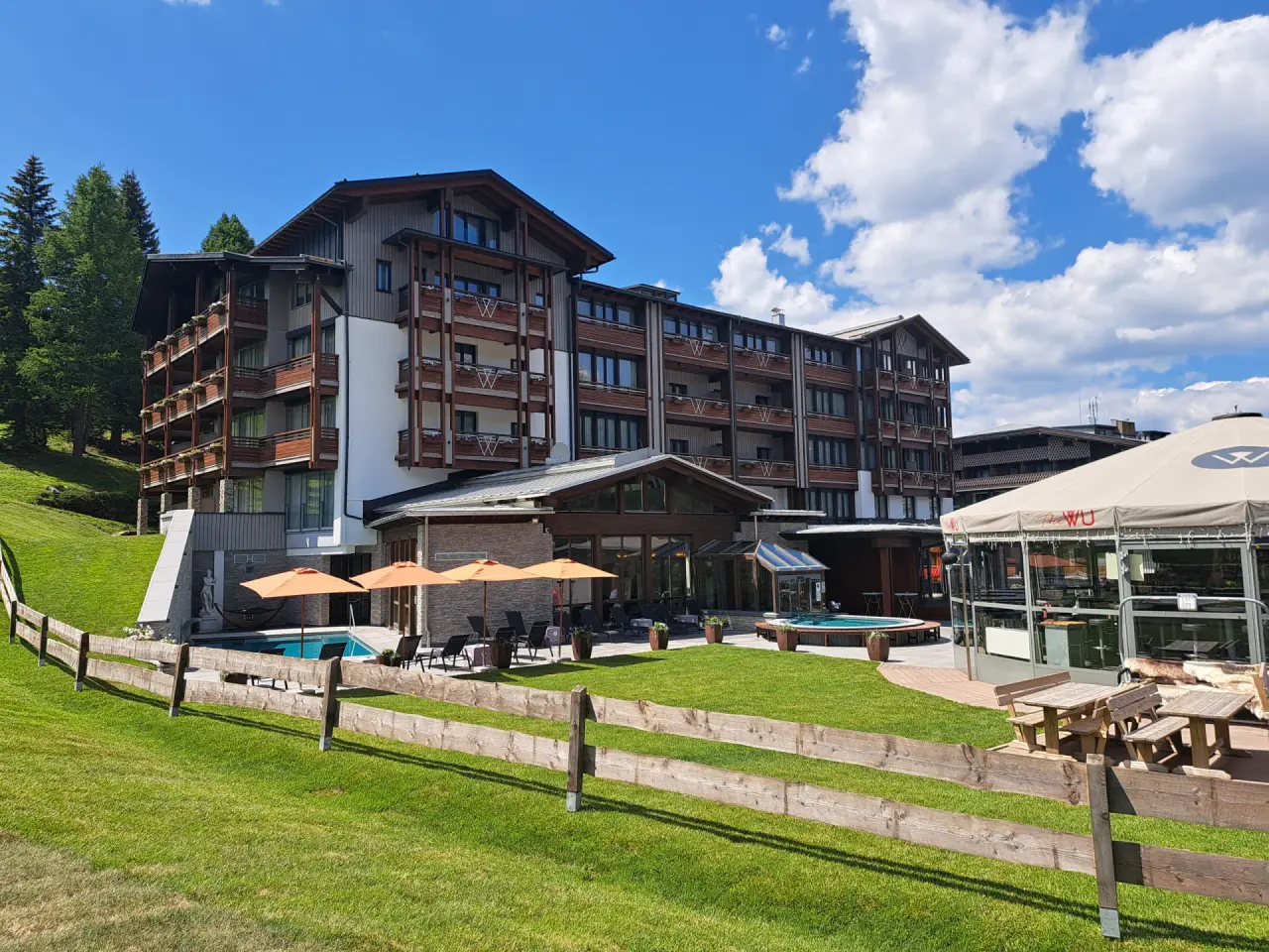 Aussenansicht des Hotel & Spa Wulfenia mit alpiner Architektur, grünem Garten, Aussenpool und Whirlpool unter blauem Himmel.