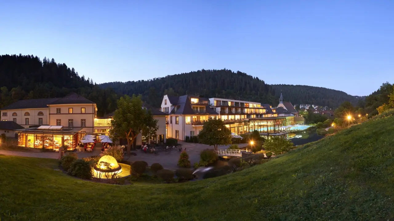 Abendlicher Panoramablick auf das Hotel Therme Bad Teinach mit beleuchtetem Gebäude und Aussenpool inmitten grüner Natur.