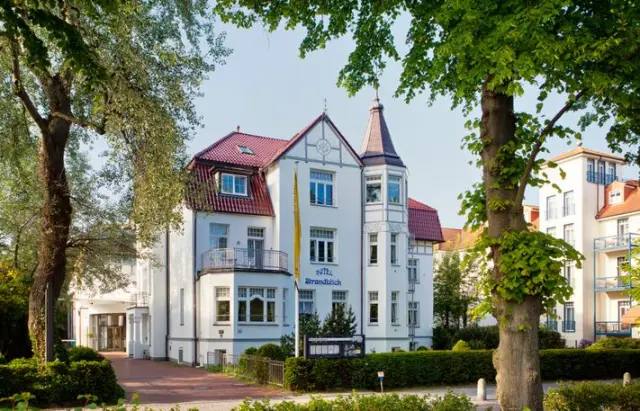 Historisches Ringhotel Strandblick mit weisser Fassade, rotem Dach und grünen Bäumen an einem sonnigen Tag.