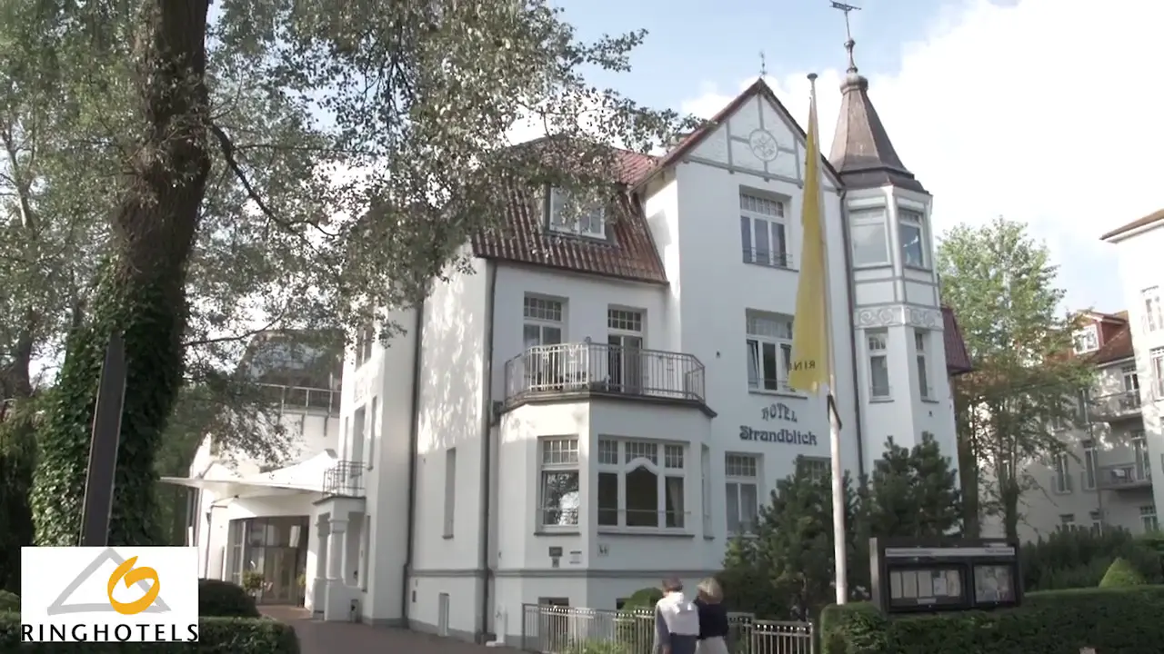 Historische Aussenansicht des Ringhotel Strandblick mit weisser Fassade, Turm und Gruenanlage an einem sonnigen Tag.