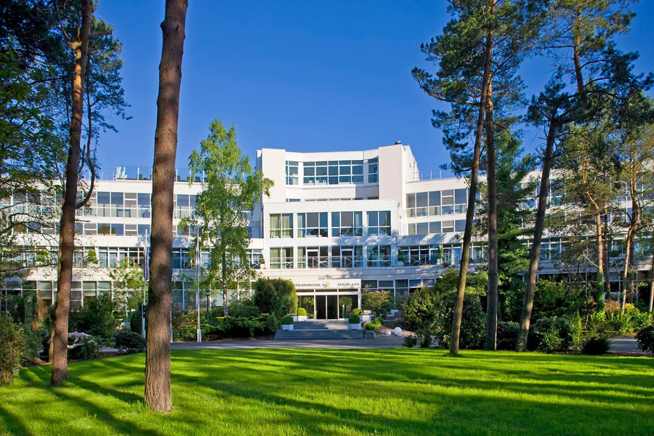 Aussenansicht des modernen Strandhotel Fischland mit gruener Wiese und hohen Baeumen unter blauem Himmel.
