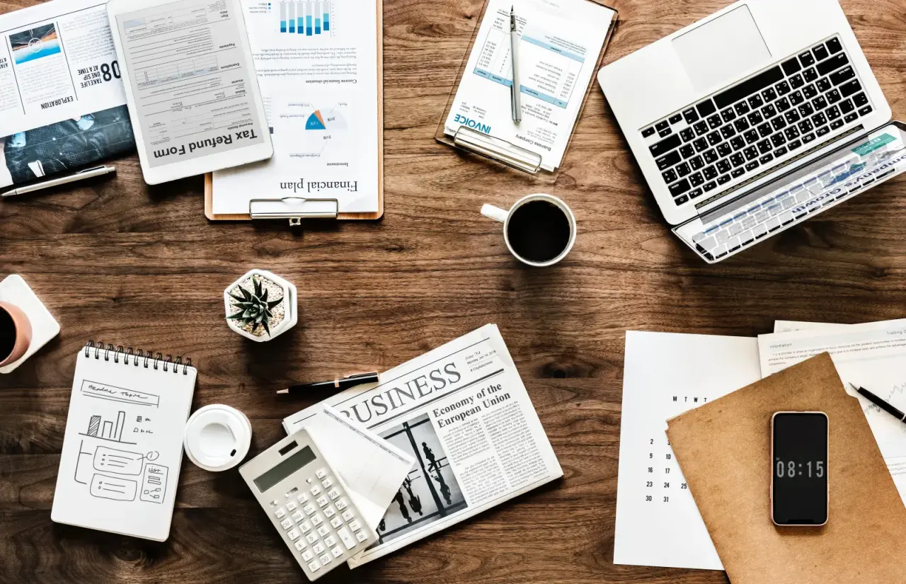 Arbeitsplatz mit Laptop, Dokumenten, Taschenrechner, Kaffee und Notizbuch auf Holztisch.