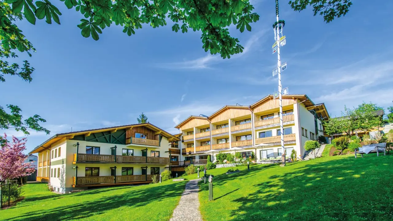 Aussenansicht des Glashotel Zwiesel mit traditioneller Holzarchitektur, grüner Gartenanlage und Maibaum bei Sonnenschein.