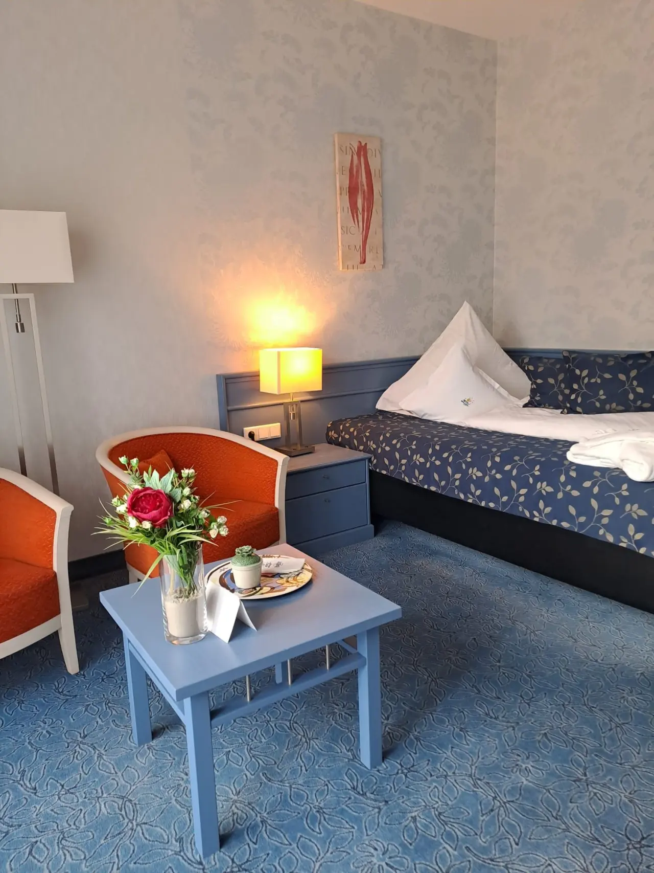 Gemütliches Hotelzimmer im Hotel Pabst mit blauem Teppich, Doppelbett, orangefarbenen Sesseln und Willkommens-Deko.