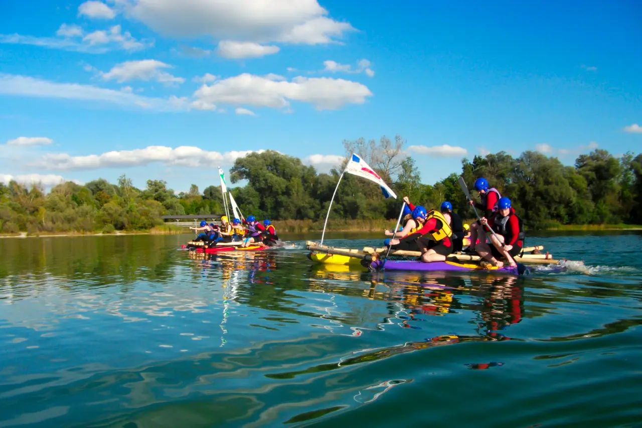 Gruppe von Personen auf selbstgebauten Flößen beim Rafting auf einem See unter blauem Himmel, Hotel Bad Schachen.