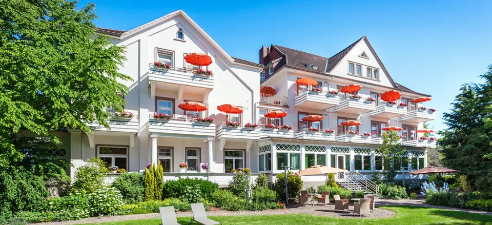 Hotel Noltmann-Peters mit charmanter weißer Fassade, roten Sonnenschirmen auf Balkonen und sonnigem Garten mit Liegen.