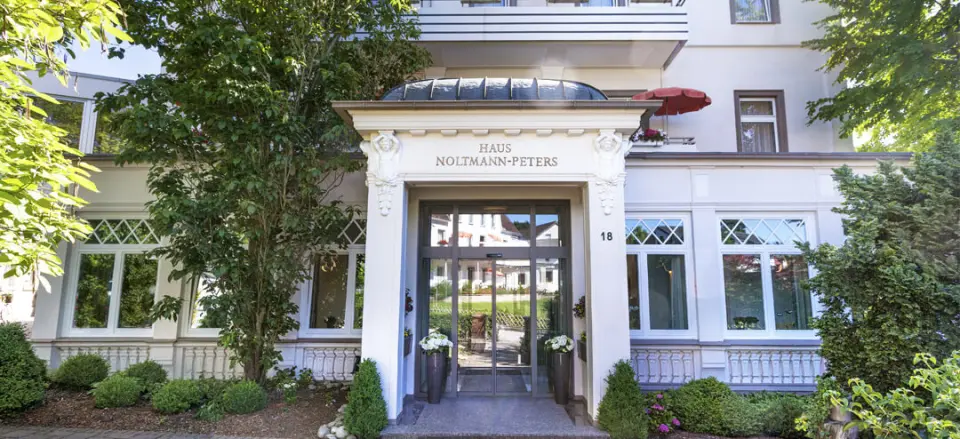 Haupteingang des Hotel Noltmann-Peters mit klassischer Architektur und grüner Bepflanzung an einem sonnigen Tag.