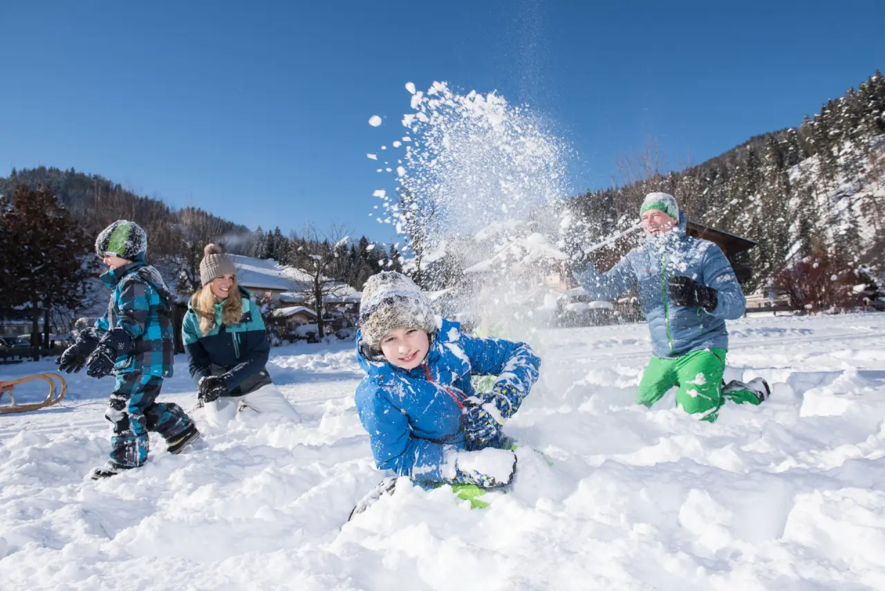 Familie spielt fröhlich im Schnee vor verschneiter Bergkulisse im Familienparadies Sporthotel Achensee.