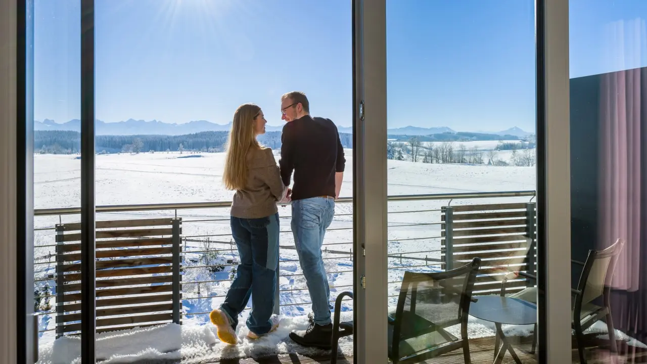 Paar auf Balkon mit Blick auf verschneite Allgäuer Landschaft und Berge im Hotel Das Weitblick Allgäu.