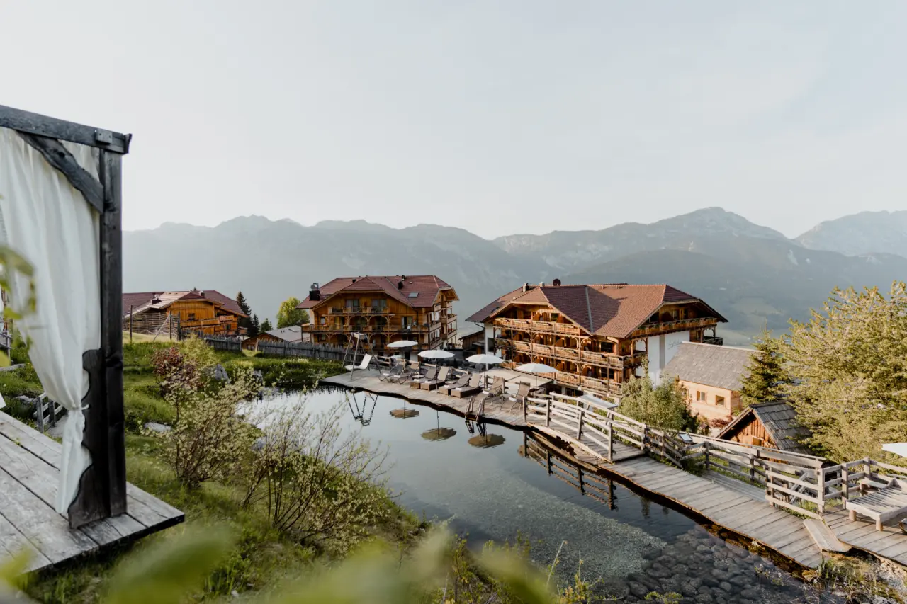 Natur- und Wellnesshotel Höflehner: Idyllischer Naturbadeteich mit Hotelanlage und majestätischem Alpenpanorama.