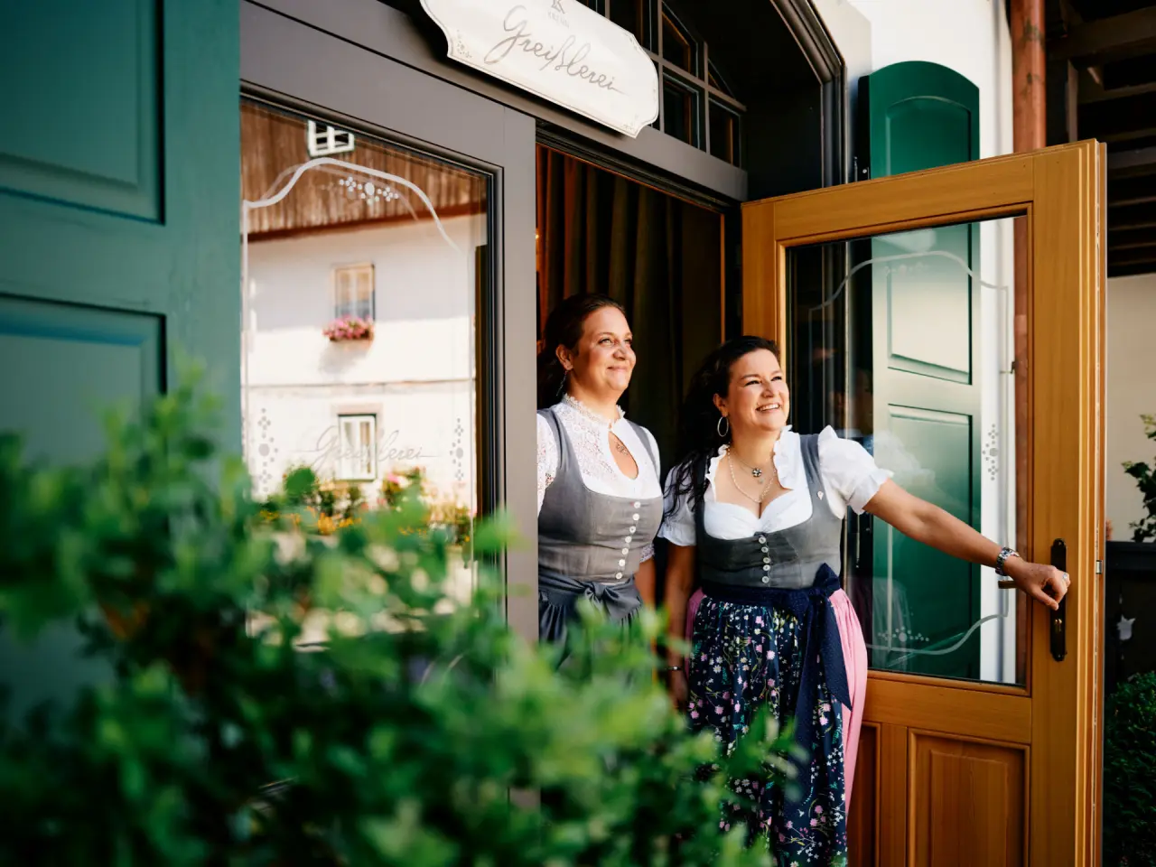 Zwei lächelnde Frauen in Tracht am einladenden Eingang der Greißlerei im G’Schlössl Murtal.