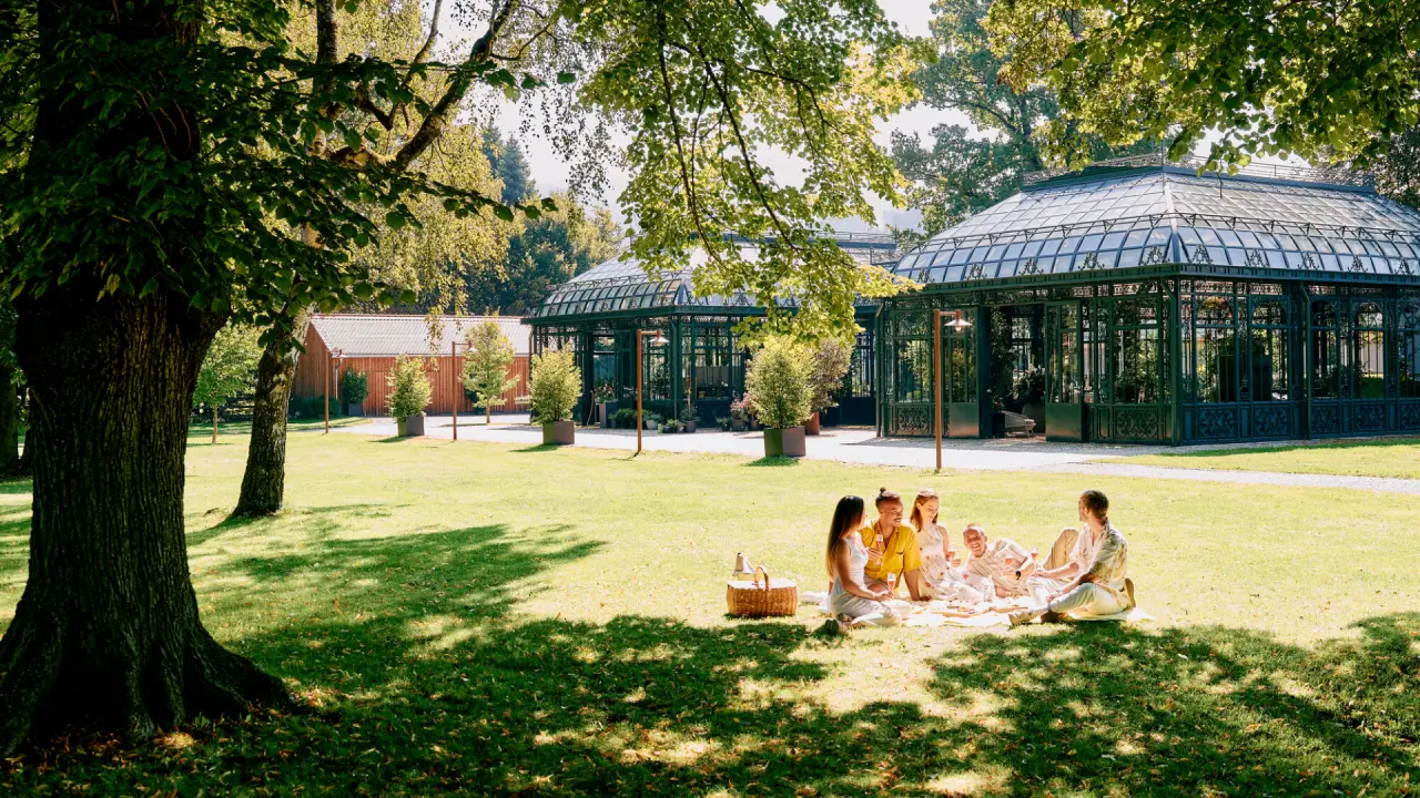 Gäste genießen ein Picknick im sonnigen Garten des G’Schlössl Murtal mit historischen Glashäusern im Hintergrund.