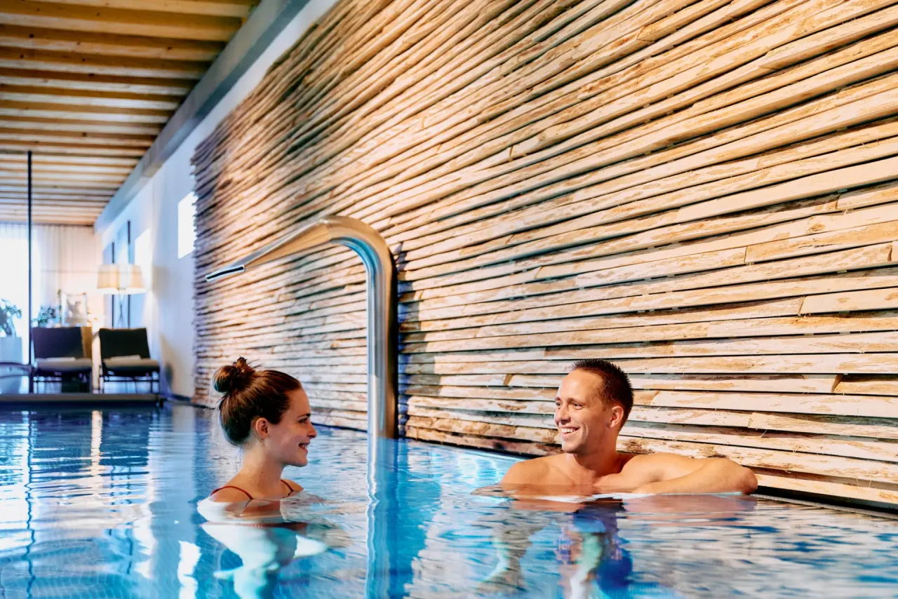 Paar entspannt im modernen Innenpool des G’Schlössl Murtal mit Holzwand und Wasserfall. Ideal für Wellnessurlaub.