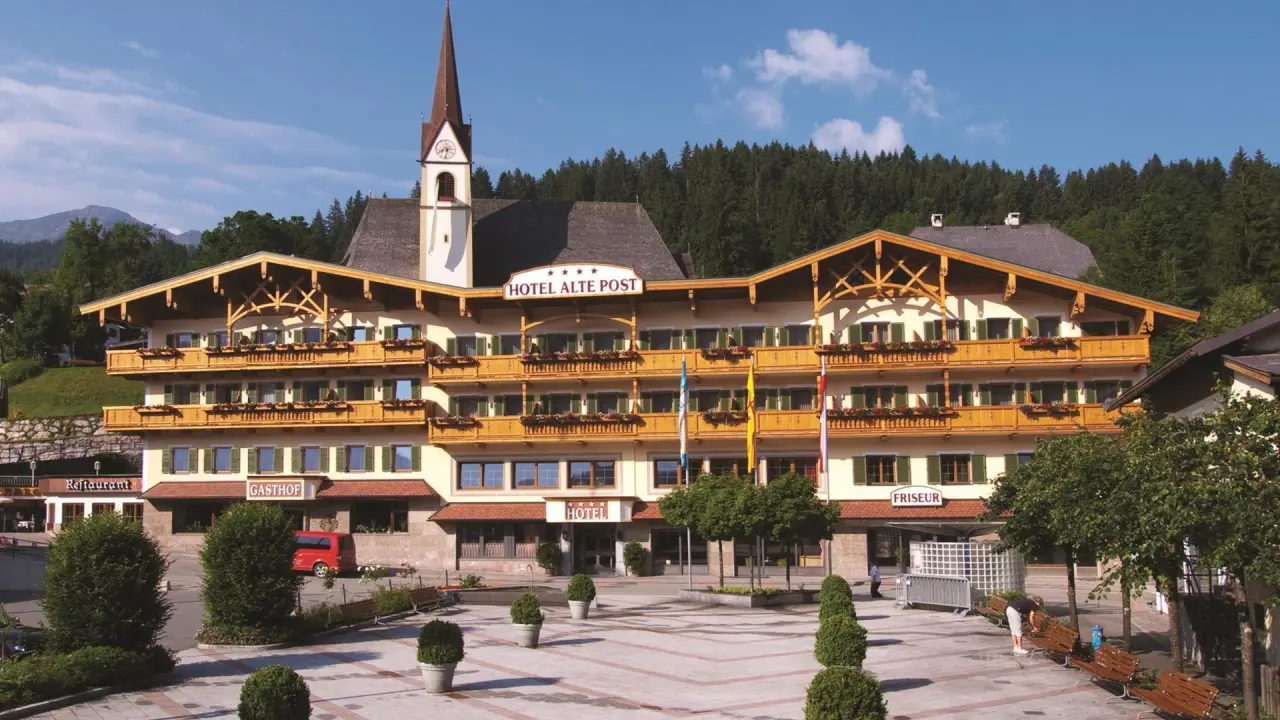 Traditionelles Hotel Alte Post in den Alpen mit Holzbalkonen und Kirchturm im Hintergrund unter blauem Himmel.