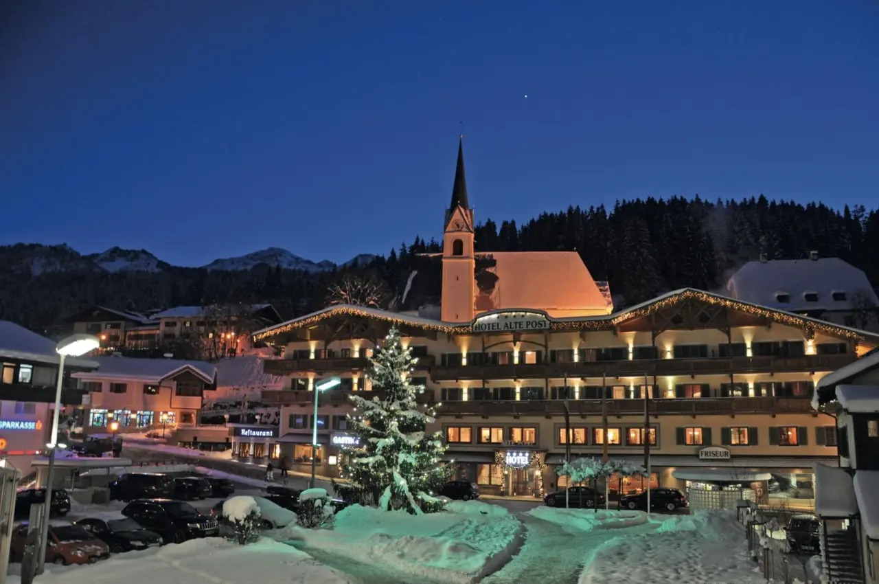 Hotel Alte Post in verschneiter Winterlandschaft bei Nacht, mit festlicher Beleuchtung und Kirche vor Bergpanorama.