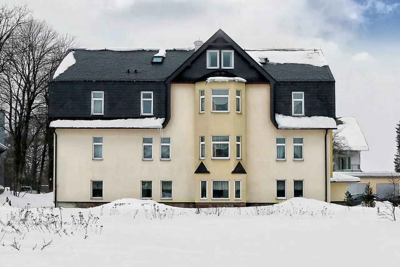 Aussenansicht des Konsum Berghotel Oberhof im Winter mit verschneitem Dach und Vorgarten unter bewölktem Himmel.