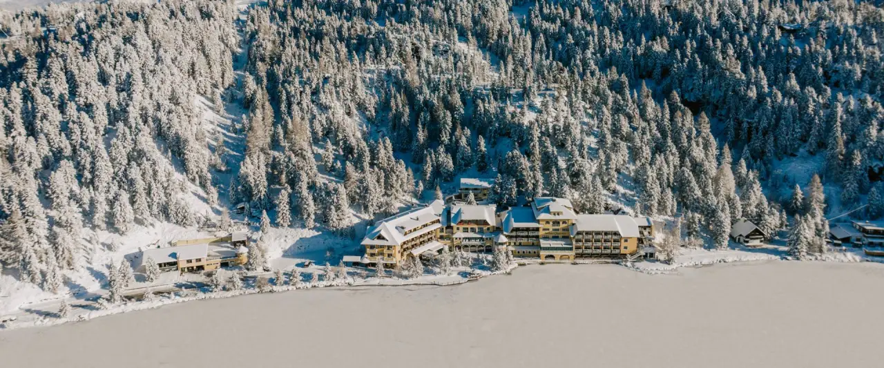 Romantik Seehotel Jägerwirt im Winter: Luftaufnahme des Hotels am zugefrorenen See, umgeben von verschneiten Wäldern.