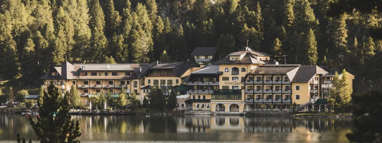 Romantik Seehotel Jägerwirt am Ufer des Aichwaldsees, umgeben von grünen Wäldern, spiegelt sich im ruhigen Wasser.
