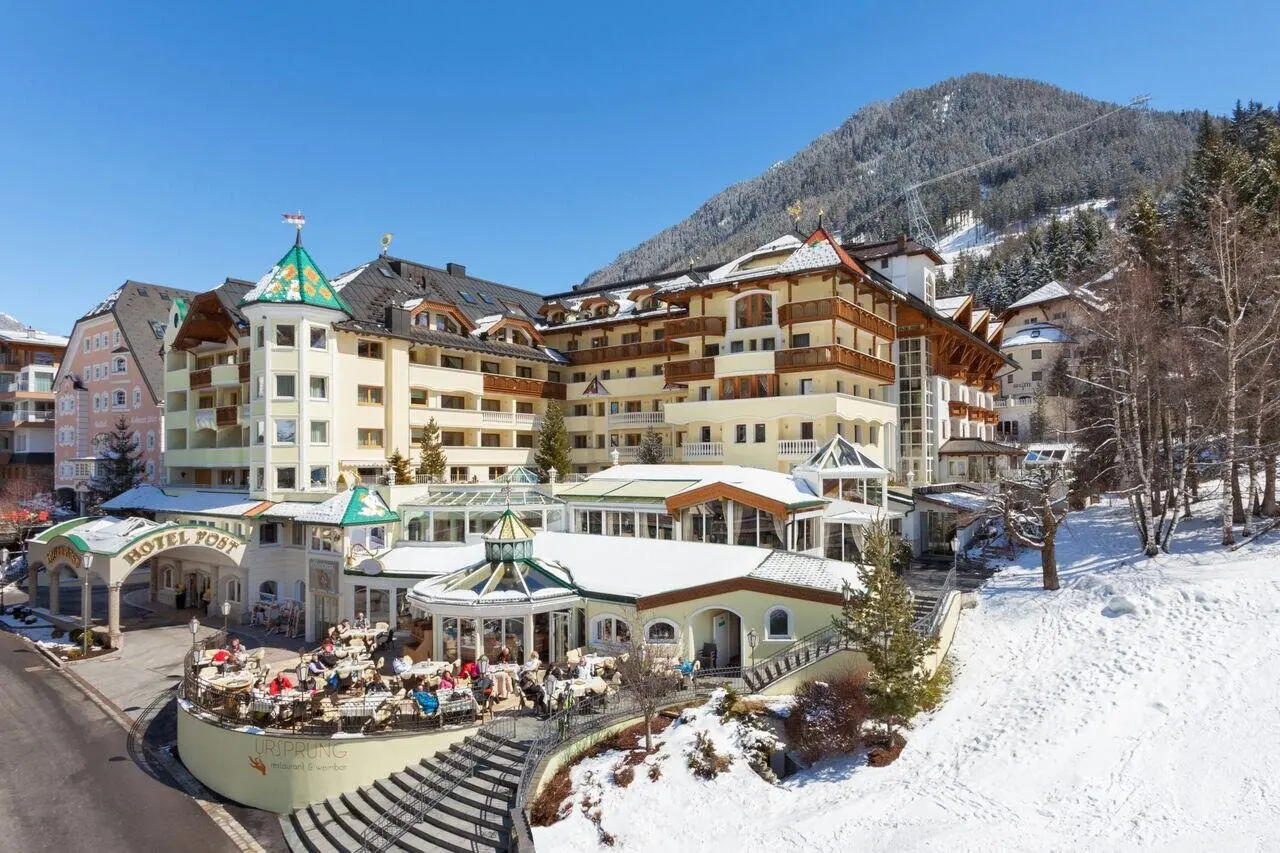 Majestätisches Hotel Post Ischgl im Winter mit schneebedeckten Dächern und belebter Sonnenterrasse vor Alpenpanorama.