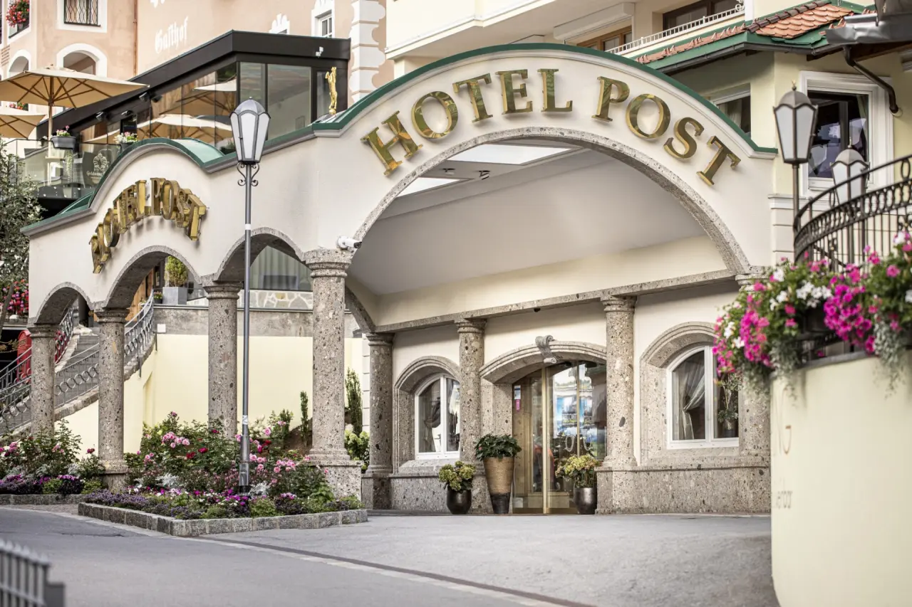 Haupteingang des Hotel Post Ischgl mit goldener Beschriftung, Steinbögen und farbenfrohen Blumenbeeten.