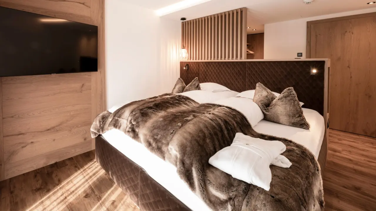 Einladendes Hotelzimmer mit Holzakzenten, großem Bett, flauschiger Decke und Bademantel im Hotel Post Ischgl.