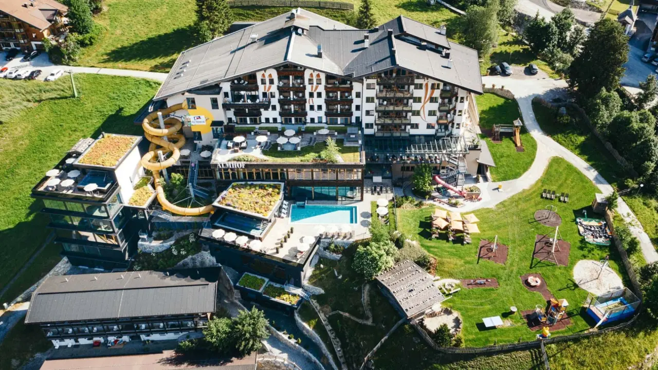 Luftaufnahme des Almhof Family Resort & SPA mit Hotelgebäude, Aussenpools, Wasserrutsche und grossem Kinderspielplatz in den Alpen.