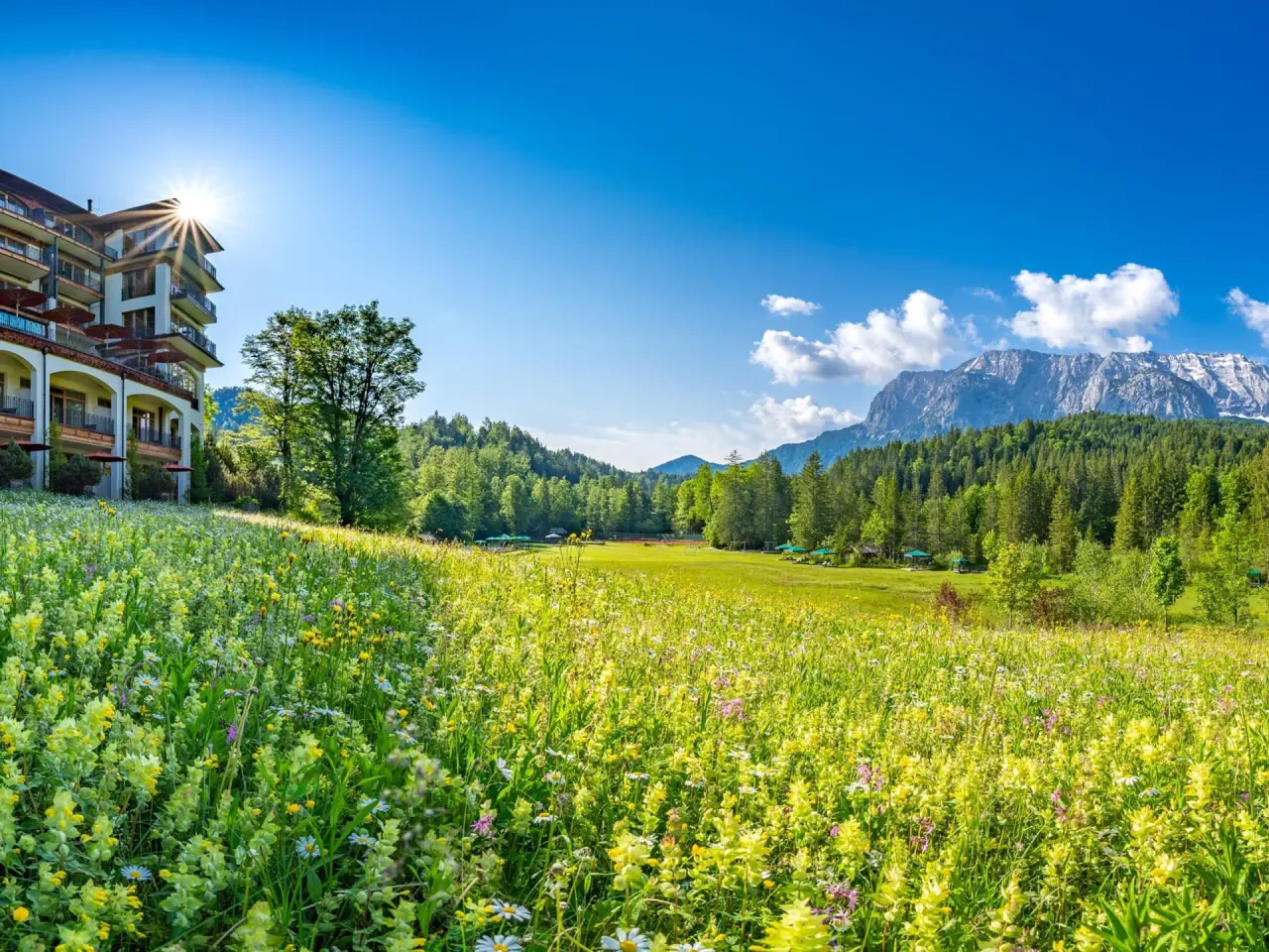 Schloss Elmau Hotel im Frühling, umgeben von blühenden Wiesen und majestätischem Bergpanorama der Alpen unter blauem Himmel.